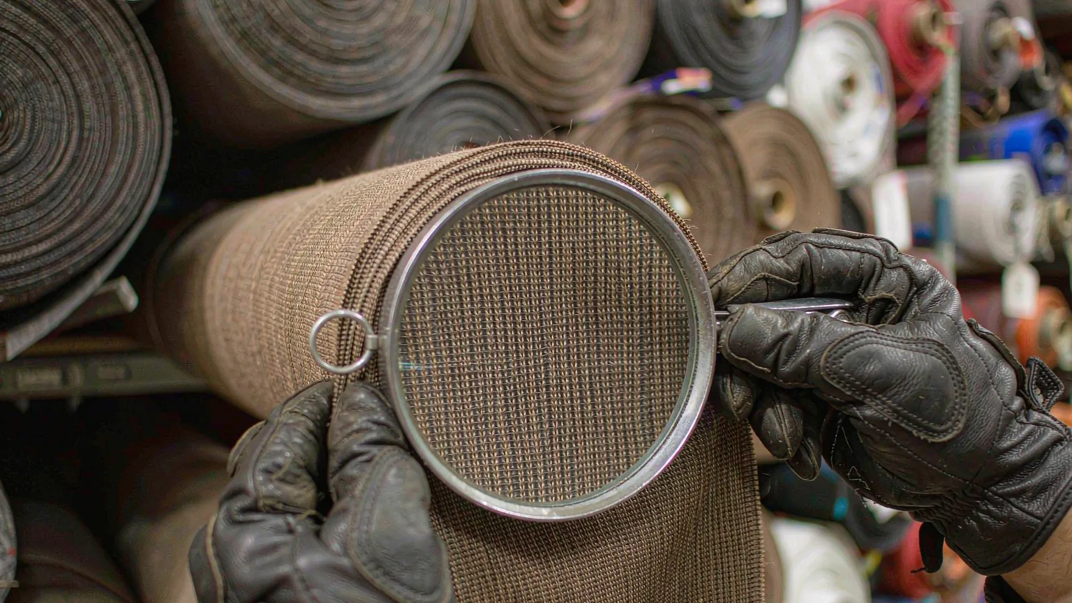 UTS inspector examining the weave of a fabric roll with a jeweller's loupe, stocked fabric rolls on the shelving behind