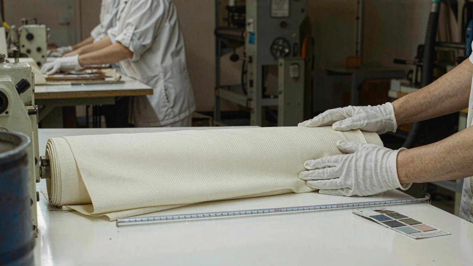 UTS inspector in white gloves unrolling cream-coloured fabric flat across the inspection table, with a tape measure and colour swatch card at the edge of the bench