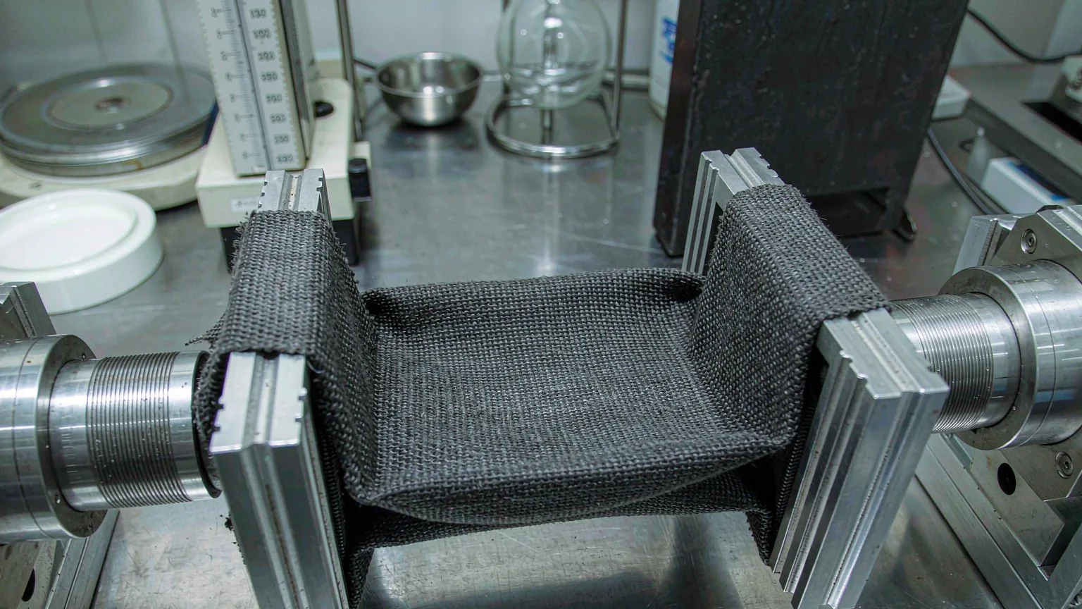 A dark woven fabric swatch clamped in a laboratory tensile testing machine, checked before bulk production runs