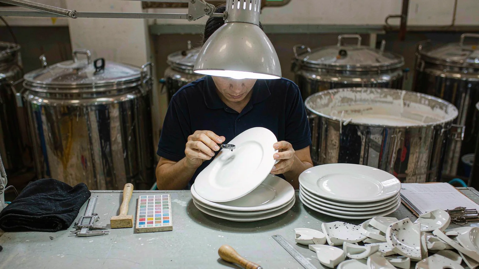UTS inspector examining the glaze of white ceramic plates under a task lamp, with rejected pieces showing pinholes, black specks and a chipped rim beside the bench