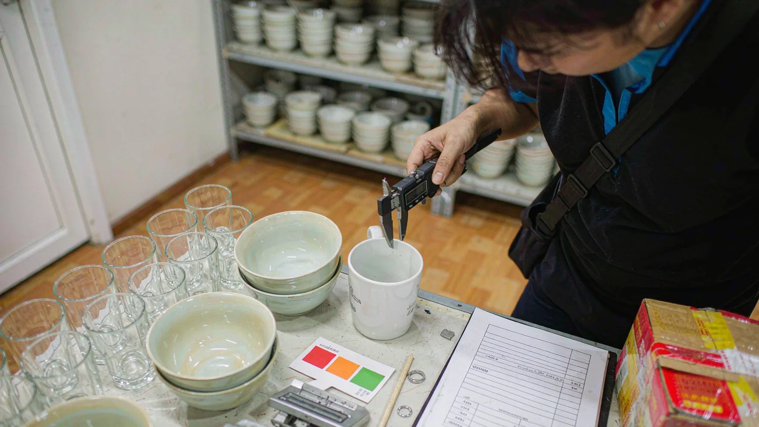 UTS inspector measuring the rim of a white ceramic mug with a digital caliper beside glass tumblers, a colour swatch card and a sealed export carton