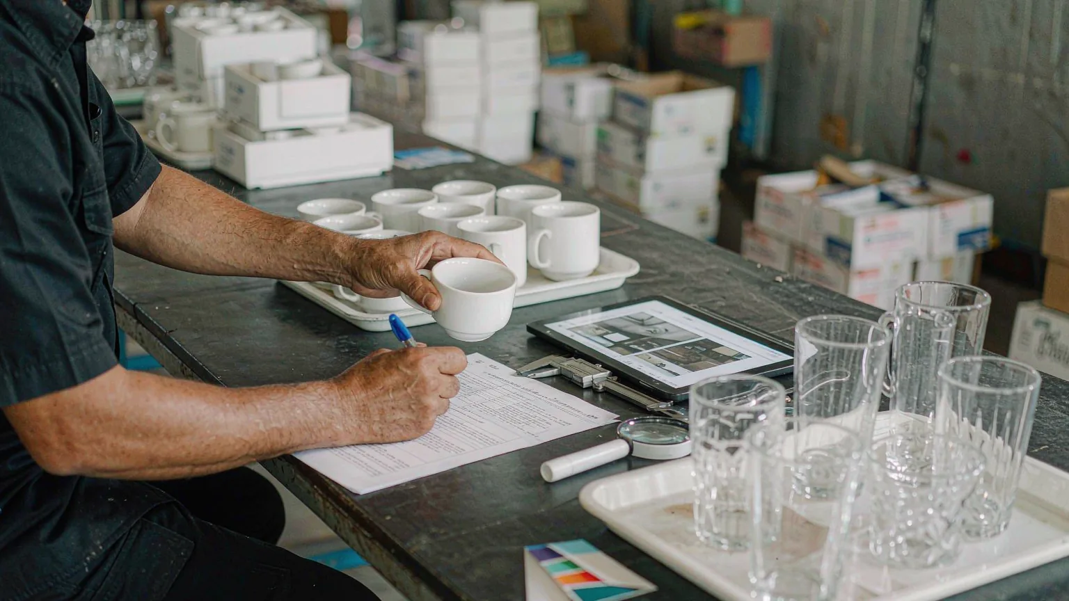 UTS inspector writing up a printed glassware and ceramic inspection report beside a tray of mugs, glass tumblers and a tablet showing defect photographs