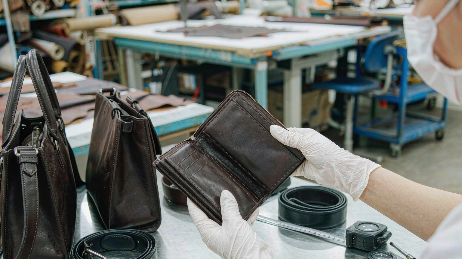 UTS inspector examining a finished dark leather wallet with gloved hands on an inspection table, with leather handbags, a tape measure and colour swatches beside
