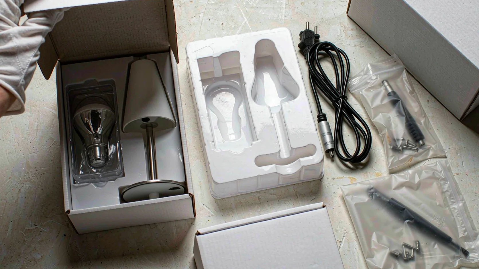 Hands unpacking a table lamp and LED bulb from the retail carton, checking the moulded inner liner, accessory bag, cable and outer shipping carton during a lighting inspection