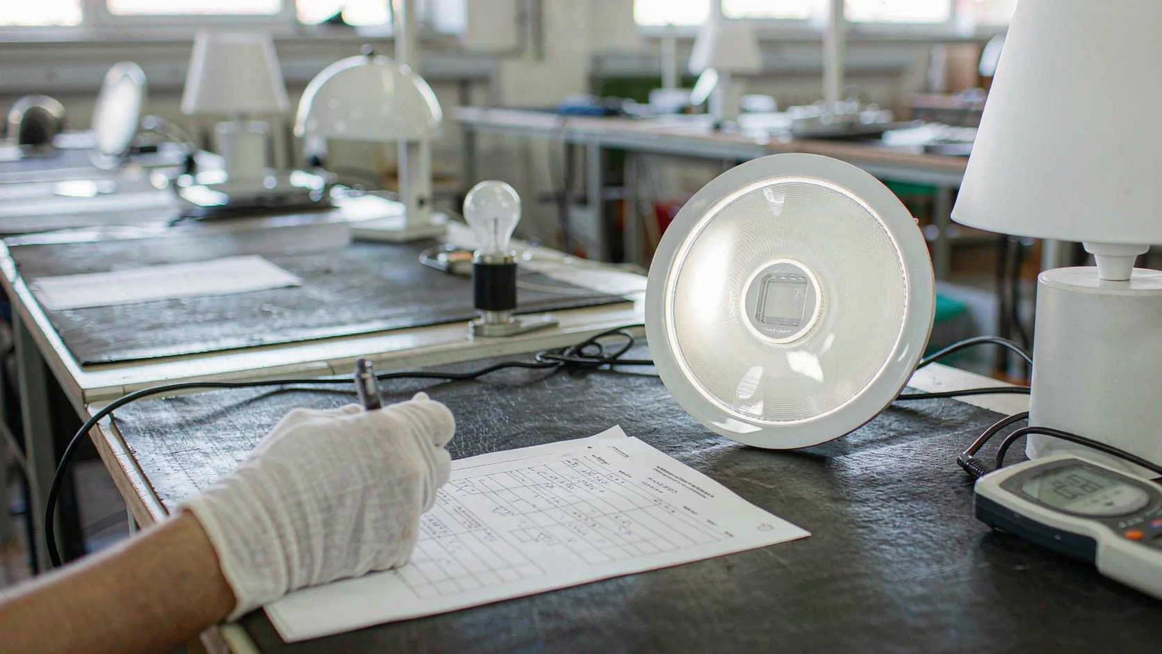 Inspector in white gloves writing measured light output data on an inspection report beside a lit LED panel, a table lamp and a lux meter on the test bench