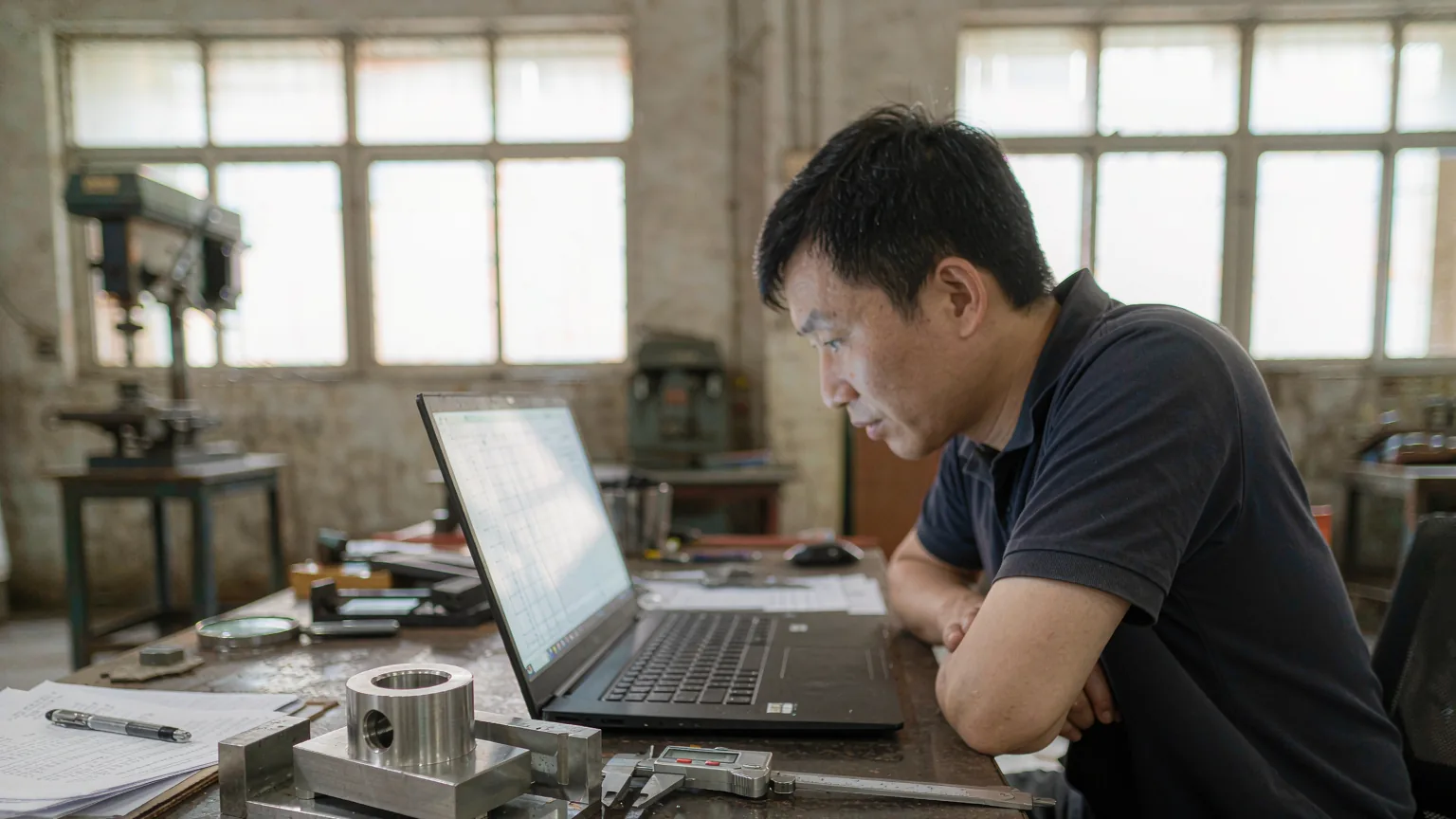 Inspector finalising the inspection report on a laptop with a caliper-measured machined part on the desk