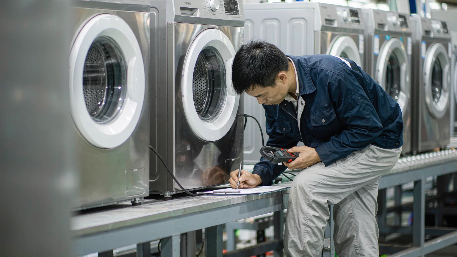 Inspector function-testing a washing machine on the factory test bench with a clamp meter and clipboard